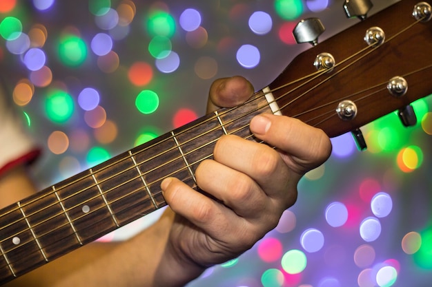 Man playing on acoustic guitar. Closeup fingers on guitar neck against christmas blurred bokeh lights on background.