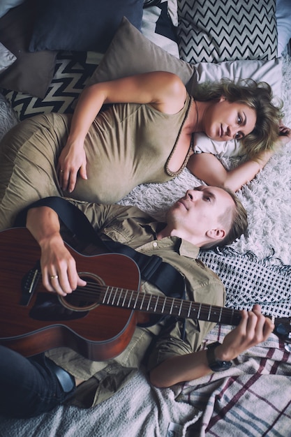 Man playing a guitar and his pregnant wife are lying on a bed in a loft room