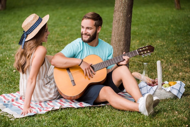 Man plays guitar for his girlfriend on the picnic in the park in the summer on sunset