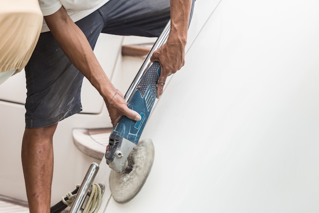  A man polishing side of the white boat by grinder machine in the marina