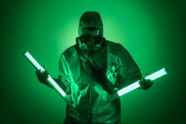 A man poses in a protective suit with a hood on his head, with a protective gas mask, posing while standing against a green background, holding uranium lamps in her divorced hands. Danger