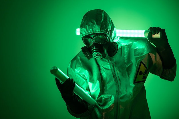 A man poses in protective suit with a hood on his head, with a protective gas mask, posing while standing on a green background, holding one uranium lamp behind his back and the other in front of him