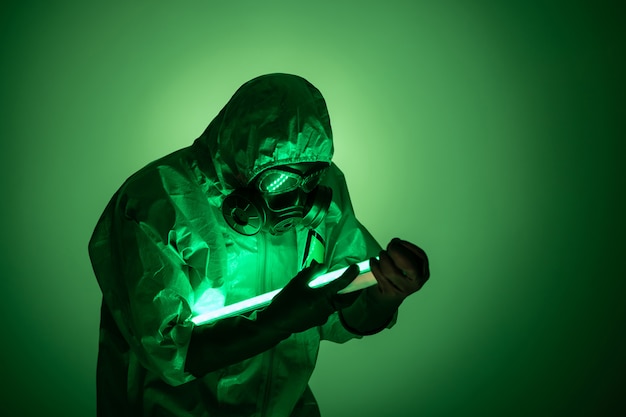 A man poses in a yellow protective suit with a hood on his head, with a protective gas mask, posing while standing against a green background, holding green uranium lamps in his hands