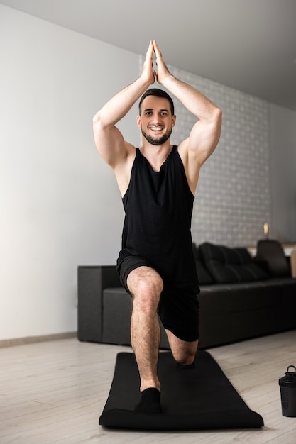 Man practicing yoga indoors doing Warrior one on black mat Muscular young man relaxing and balancing in asana Morning excercising Healthy mind in strong body concept Cozy living room on background