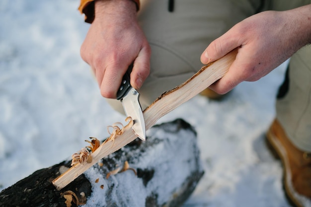 A man prepares wood wool with a knife for starting a fire in a winter forest at sunset Winter survival concept