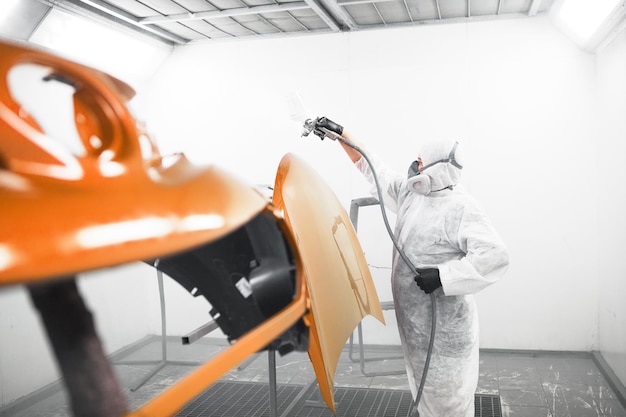 Man in protective mask and clothes sprays varnish to car hood with a spray gun in a paint booth