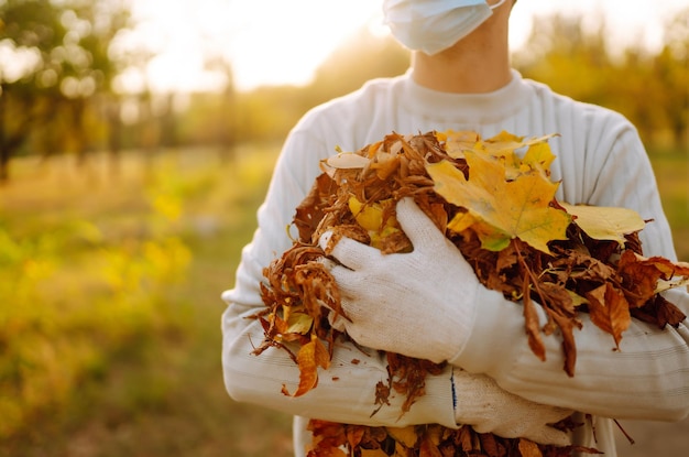 Man in protective medical mask cleans autumn leaves in the park Man in gloves collects