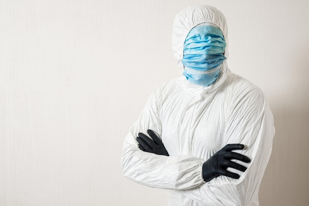 A man in a protective suit hung with medical masks posing against a wall background showing various gestures with his fingers arms folded on chest