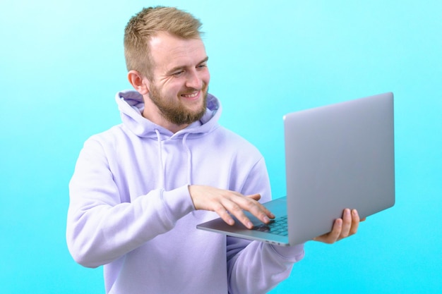 A man in a purple hoodie smiling and looking at a laptop while he typing some text on a blue background Cheerful Sign Approval Confident Corporate Good Handsome Joy Positive Successful