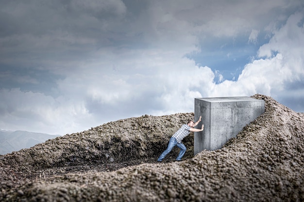 Photo man pushes a huge concrete cube crawling into the ground. concept of determination and strength.