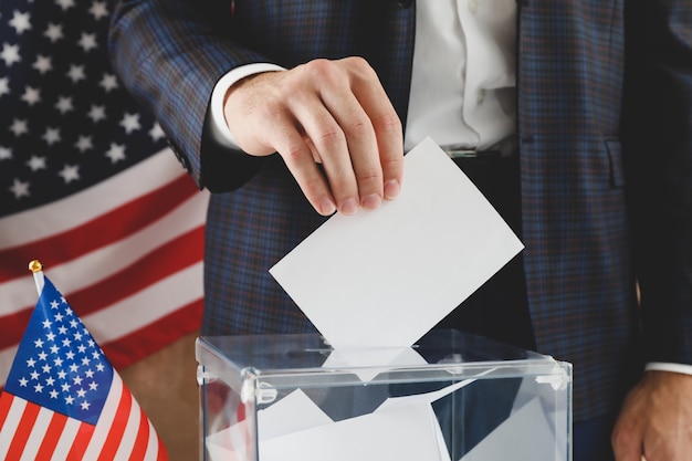 Man putting ballot into voting box against brown surface with american flag