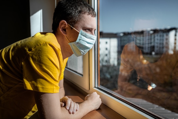 Photo man in quarantine because of a virus sits at home in a mask and looks out the window on a sunny day.
coronavirus concept. covid-19