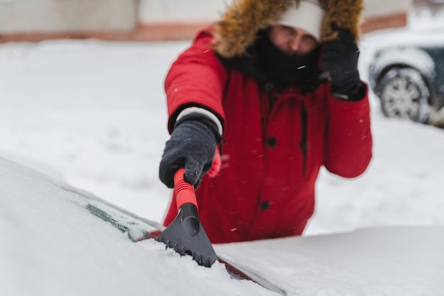 Man in red winter coat with fur hood cleaning car after snow storm