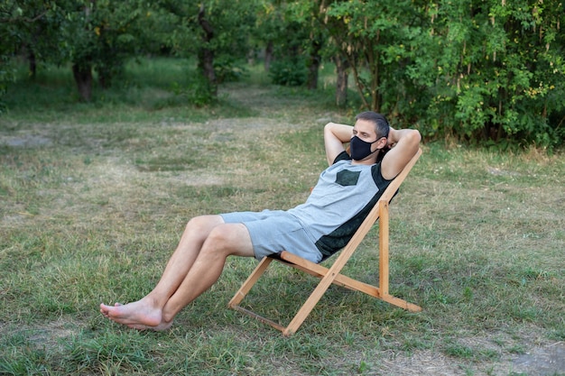 Man relaxing on deck chair in protective medical mask on his face sunbathing outside green lawn in park