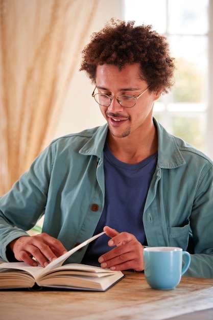 Man Relaxing At Home Sitting At Table Reading Book With Hot Drink