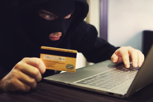 Photo man in robber mask and hood misappropriates personal bank data cyber fraudster attacks online banking system hacker makes financial fraud via internet digital crime with the use of payment card