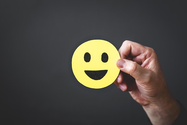 Man's hand in a business suit holds a yellow smiley face with a happy face icon on a dark background The concept of customer satisfaction or positive feedback