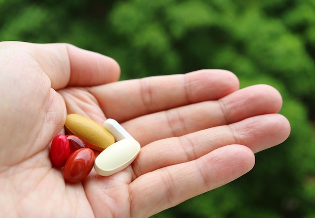 Man's Hand Holding Assorted Supplement Pills Against Blurry Green Foliage