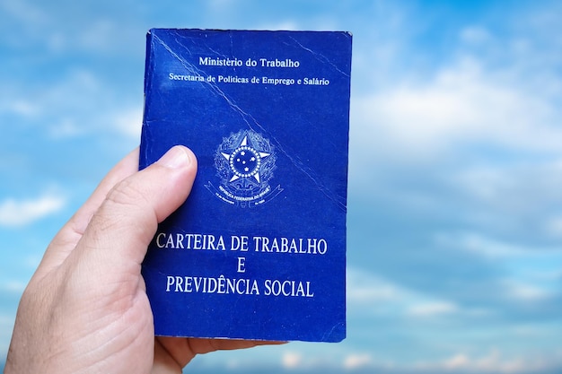 Man's hand holding Brazilian Work Card isolated on background with blue sky Written in Portuguese Federative Republic of Brazil Ministry of Labor Work and Social Security card Brazilian work concept