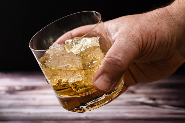 A man's hand holding a glass with whiskey or scotch and large pieces of ice from a rustic wooden table