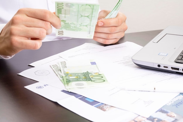 A man's hand holds out one hundred euro bills against the background of an office table with a computer