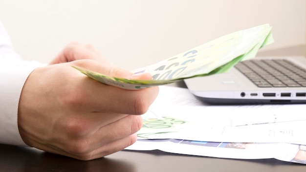 A man's hand holds out one hundred euro bills against the background of an office table with a computer