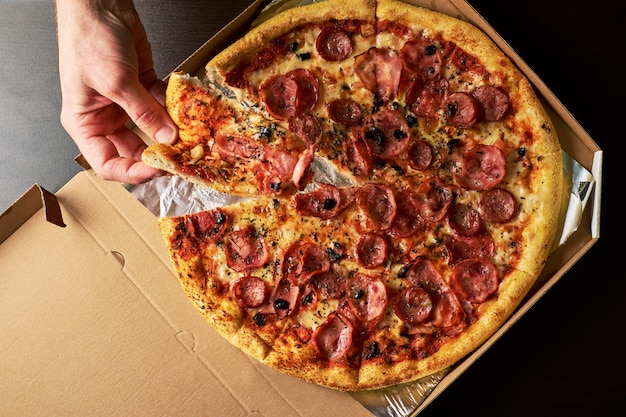 Man's hand is holding slice of cheese pizza from whole Pizza in cardboard box dark table background