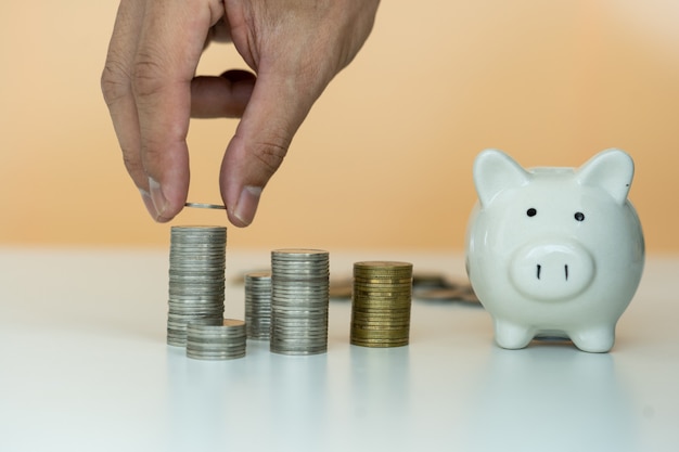 A man's hand is placing a silver coin stacked side by side with a white piggy bank. Man to collect savings as a business capital or savings money for the future. Concept saving money