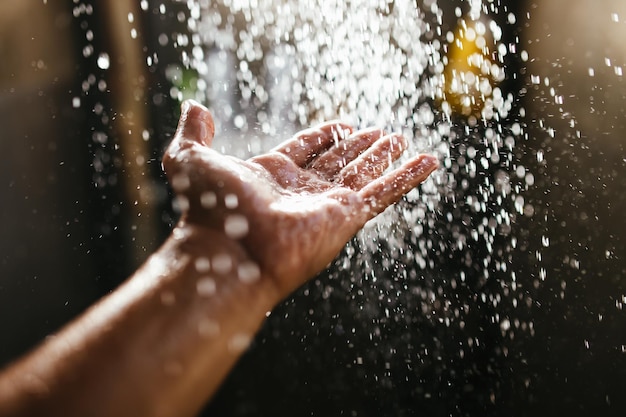 A man's hand in a spray of water in the sunlight against a dark background.