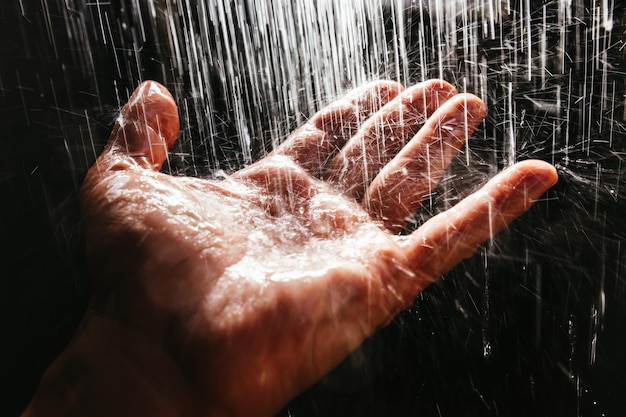 A man's hand in a spray of water in the sunlight against a dark background.