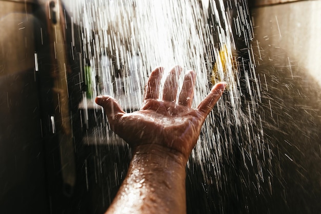 A man's hand in a spray of water in the sunlight against a dark background.