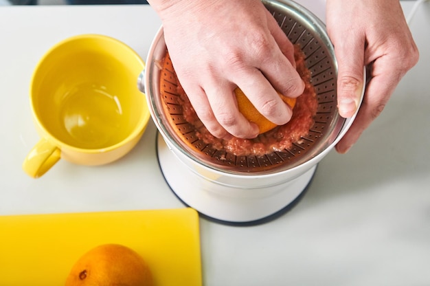 man's hand squeezes grapefruit juice on white juicer kitchen utensils and fruits on table top view