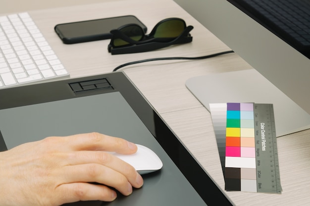 Man's hand using mouse, with colored patches, glasses and smartphone on work table