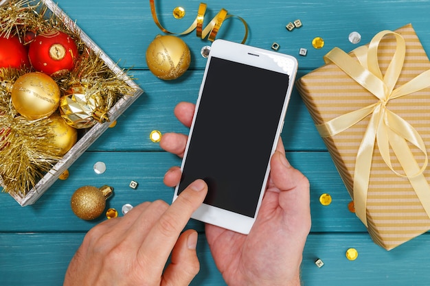 Man's hands holding a smartphone over Christmas decorations and gift box