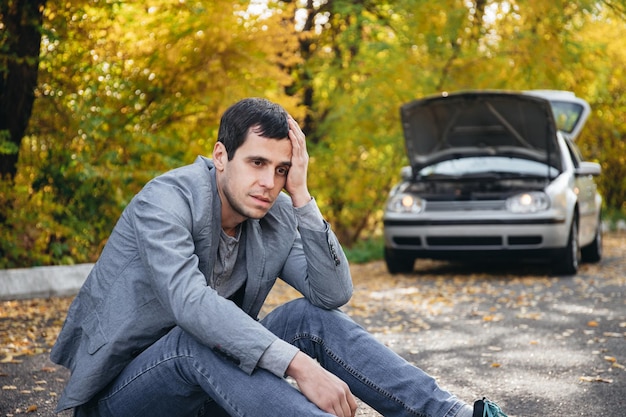 A man in sadness sits on the road in front of a broken car with an open hood