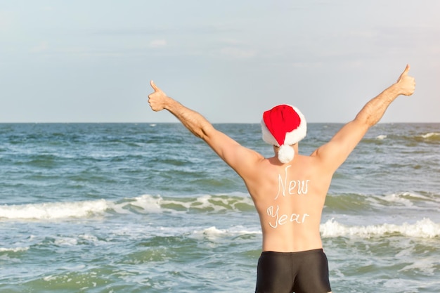 Man in Santa hats with the inscription New Year on the back on the beach Thumbs up Back view