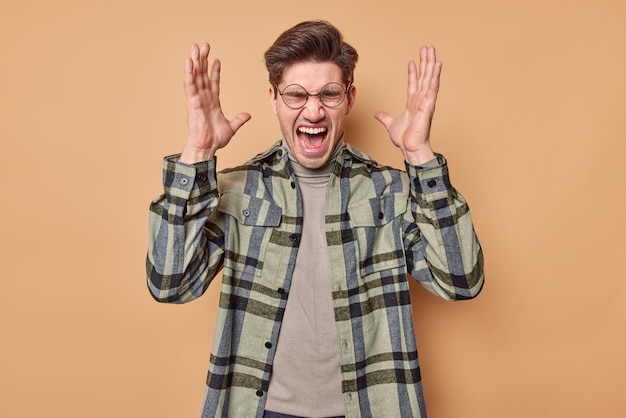 man screams loudly keeps mouth widely opened raises palms on head reacts emotionally on something wears round spectacles and checkered shirt poses on beige wall