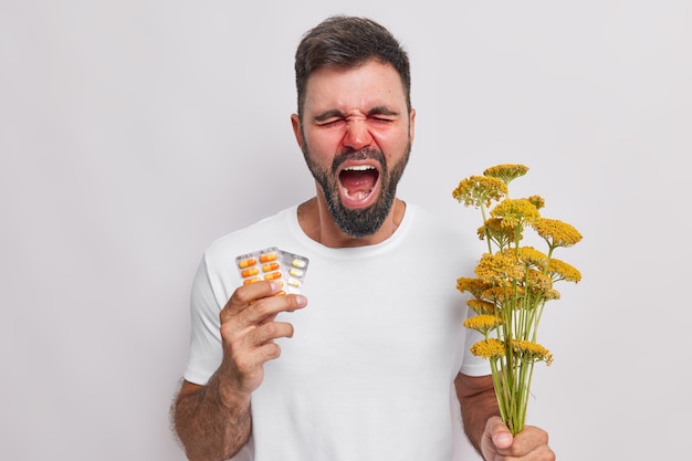  man screams loudly suffers from seasonal allergy holds medicine and bouquet of wildflowers has stuffy nose needs good treatment poses indoor on white 