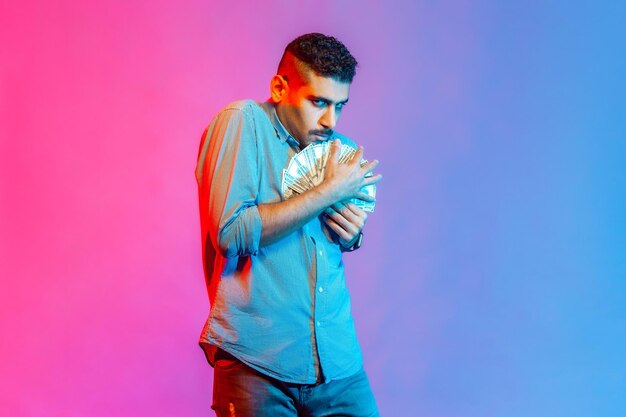 Man in shirt embracing dollar banknotes looking at camera with greedy expression, afraid to lose money, obsessed with finances. Indoor studio shot isolated on colorful neon light background.