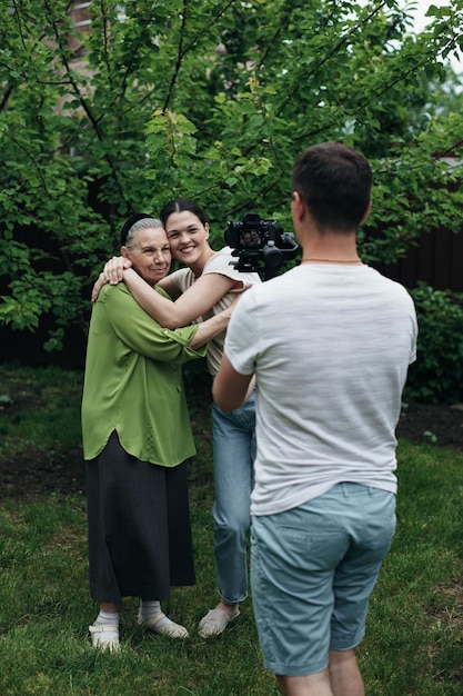 A man shoots a video on a camera as a grandmother and granddaughter hugging in the garden