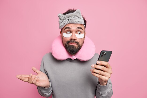  man shrugs shoulders holds mobile phone checks newsfeed wears blindfold neck pillow patches under eyes 