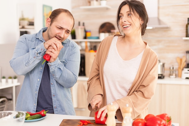 Man singing on red pepper in kitchen making wife smile. Girlfriend preparing salad. Funny happy in love couple at home spending time together, cooking healthy and smiling