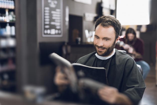 A man sits in a barbershop and selects a haircut in catalog.