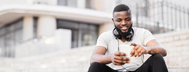 A man sits on a set of stairs while looking at his phone he is smiling and wearing headphones around