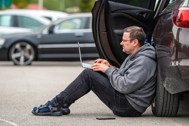 Man sitting on asphalt by car and working on laptop working remotely traveling