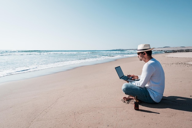 Photo man sitting on the beach with a laptop alone doing telecommuting or remote work and drinking beer