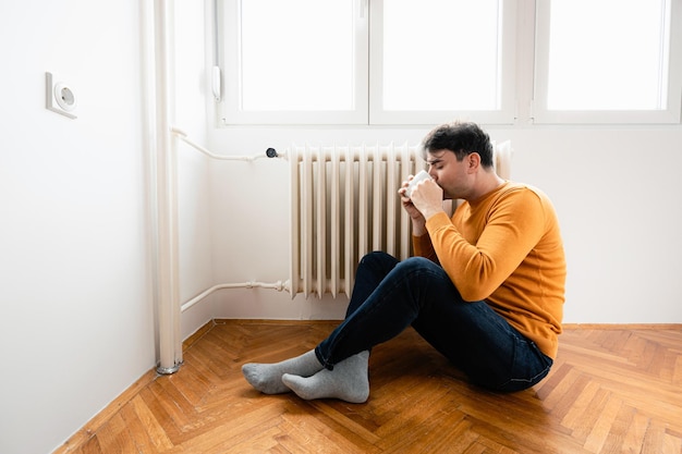 Photo man sitting by radiator drinking coffee in a room with wood floor