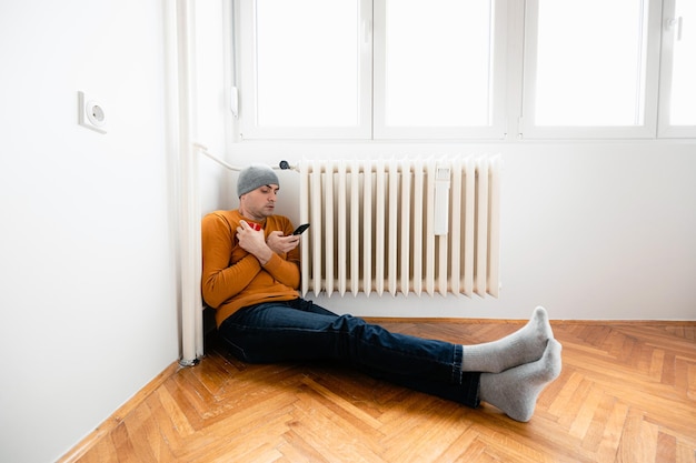 Photo man sitting by radiator with phone and mug on cold day