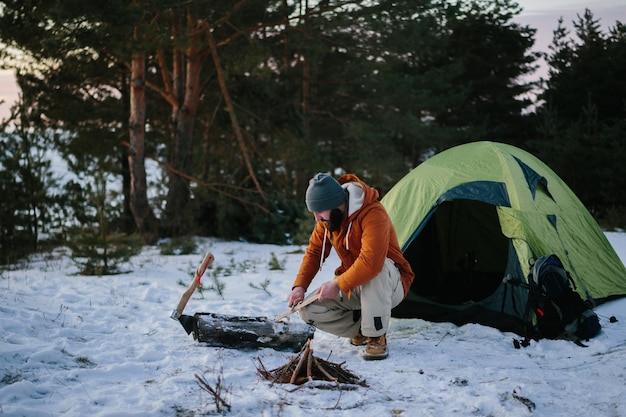 A man sitting near his tent prepares wood wool with a knife for starting a fire in a winter forest at sunset Winter survival concept