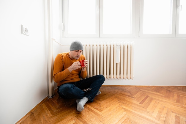 Photo man sitting near radiator in room with wood floor drinking from red mug